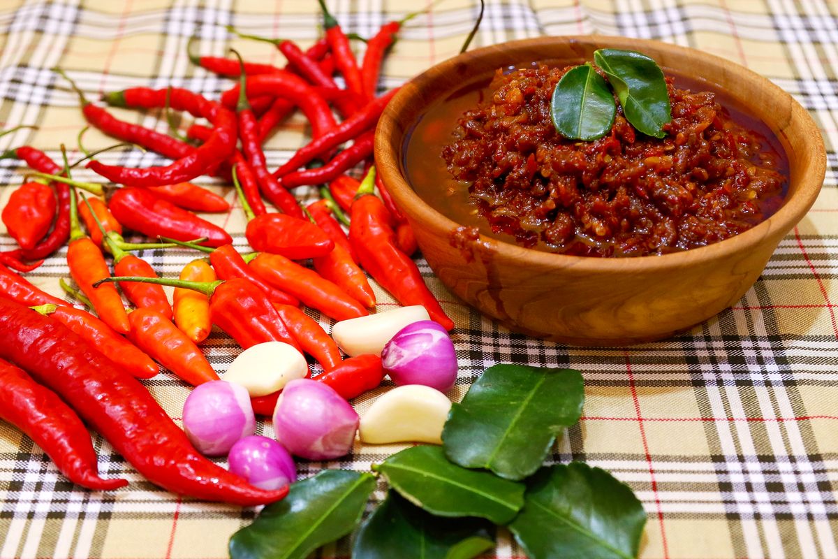 A wooden bowl of dark red chilli paste beside fresh red chillies, garlic cloves, shallots and lime leaves